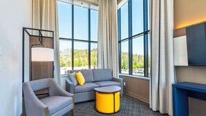 Una habitación de hotel moderna con un sofá gris, un sillón a juego, una mesita de noche amarilla y un gran ventanal con vistas a zonas verdes.