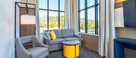 Une chambre d’hôtel moderne avec un canapé gris, un fauteuil assorti, une petite table jaune et une grande fenêtre donnant sur un paysage verdoyant.