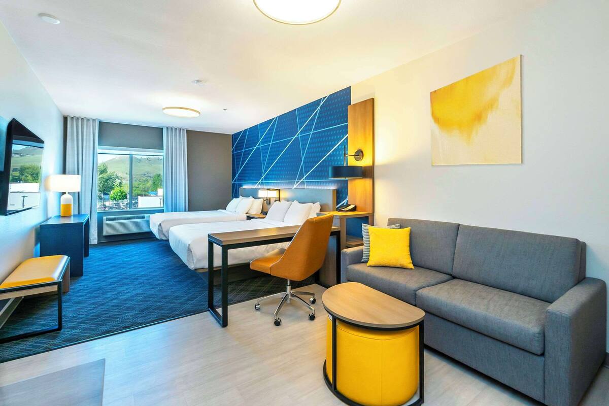 A modern hotel room with a bed, a sofa, a desk, and a chair. There is a large window with a view of the outdoors.