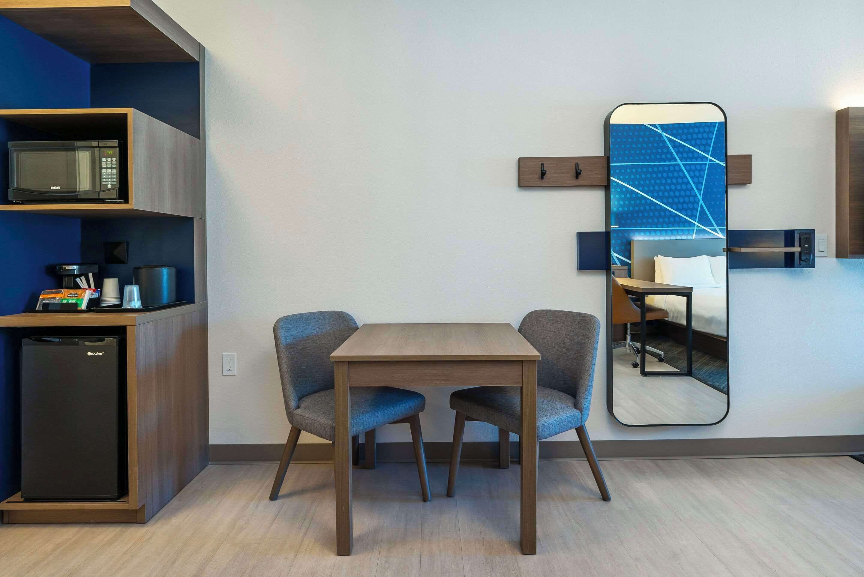 Une chambre d’hôtel compacte dotée d’une kitchenette, d’un coin repas avec deux chaises, d’un grand miroir et d’un lit avec une tête de lit.
