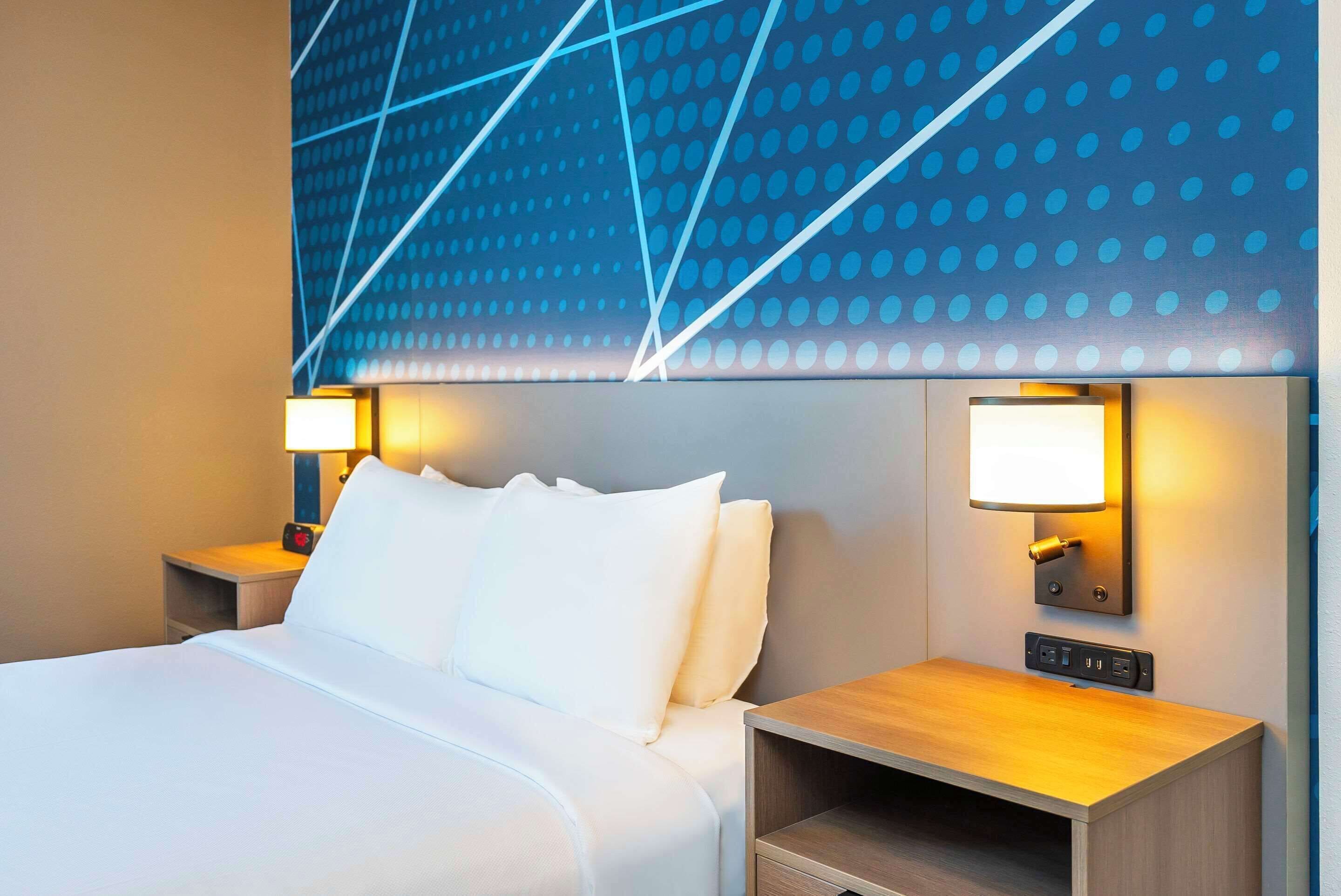 Una habitación de hotel con una cama grande, dos mesitas de noche con lámparas, una luz empotrada en la pared y un enchufe.