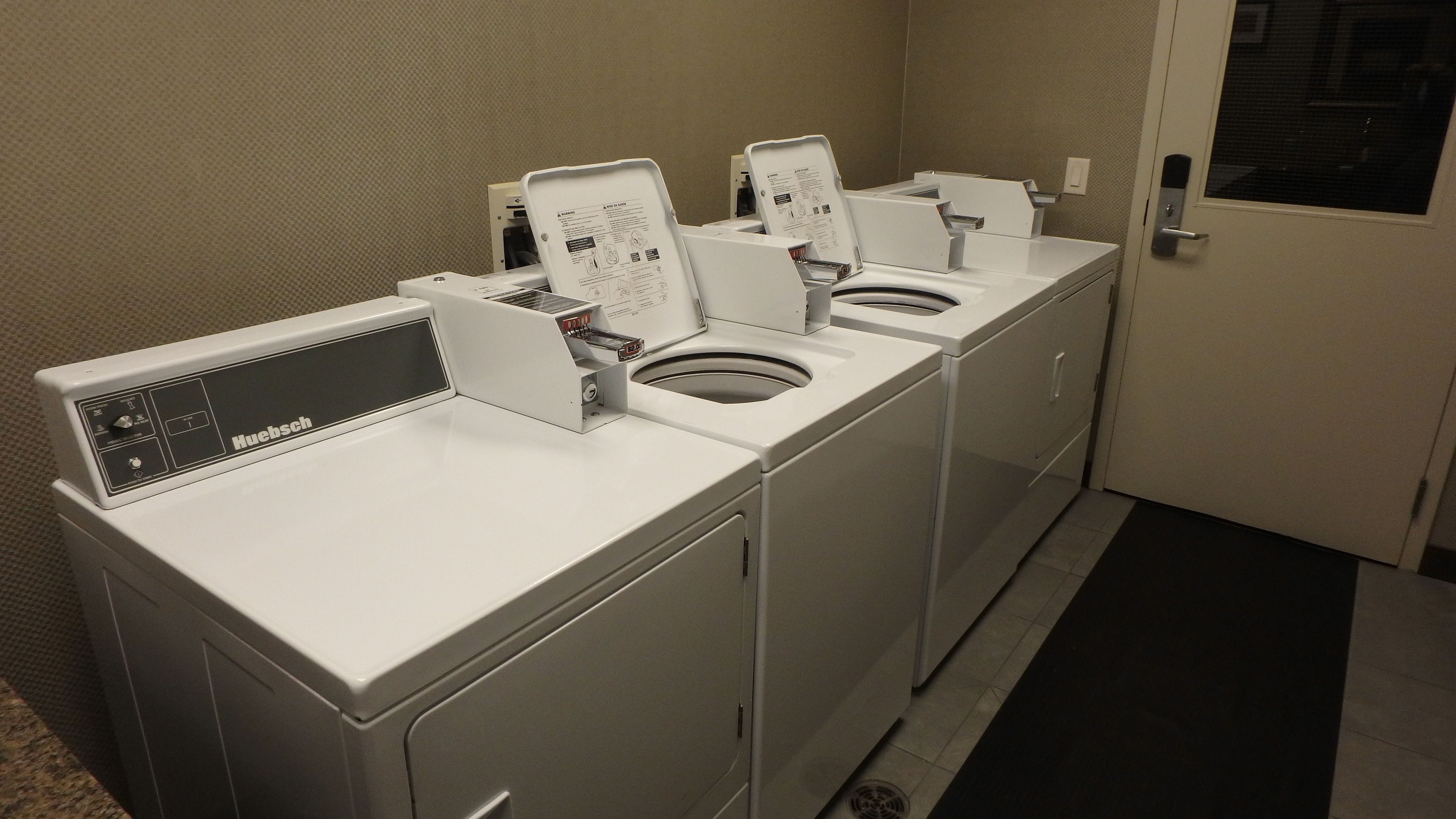 A row of white Huebsch washing machines in a laundry room with a beige wall and a door on the right.