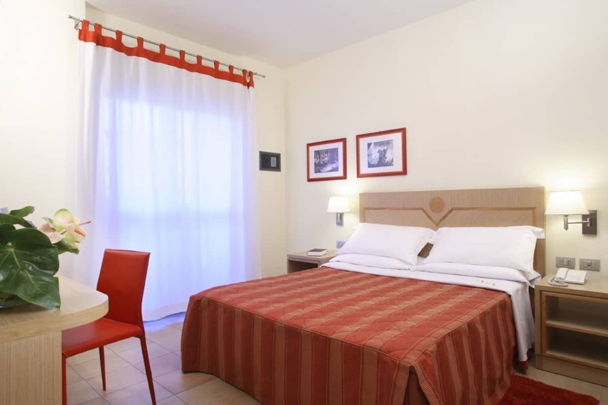 A hotel room with a bed, a desk, a chair, a nightstand, and two framed pictures on the wall.