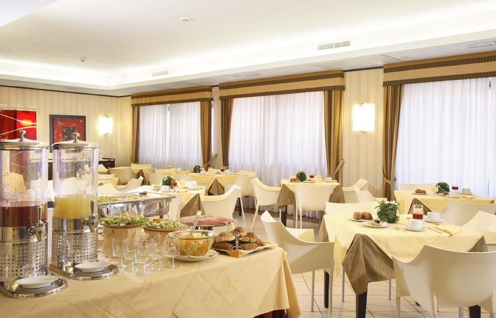 Hotellin ruokailutila, jossa on useita aamiaista varten katettuja pöytiä sekä monipuolinen buffet, jossa on tarjolla erilaisia ruokia ja juomia.
