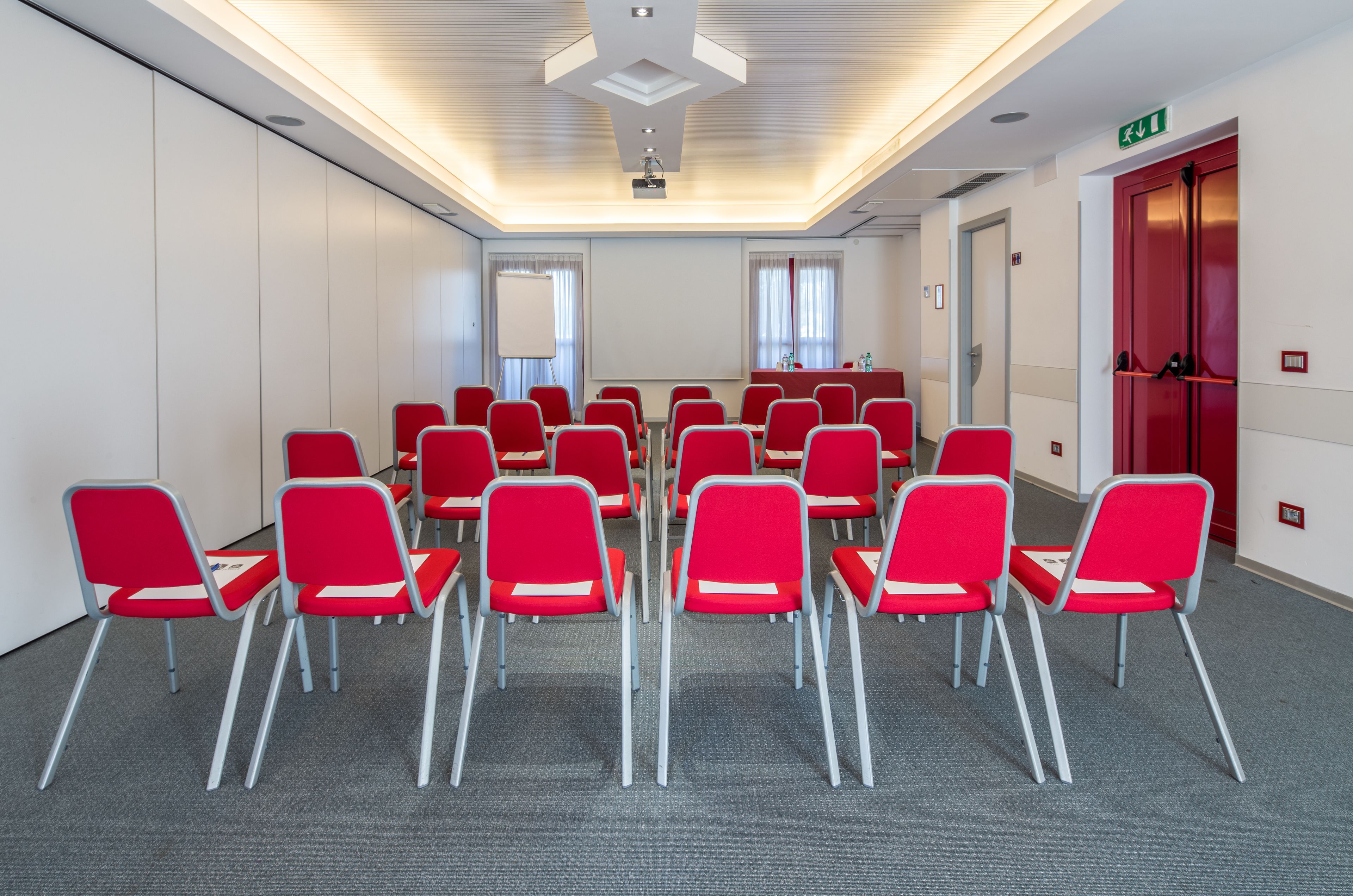 Ein Konferenzraum mit Reihen roter Stühle, einer Projektionsleinwand und einer Wandtafel.