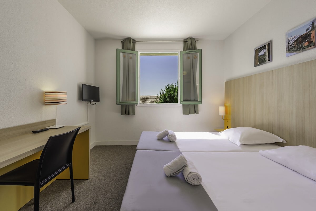 Ein Hotelzimmer mit zwei Betten, einem Schreibtisch, einem Stuhl, einem Fernseher und einem Fenster mit Vorhängen.