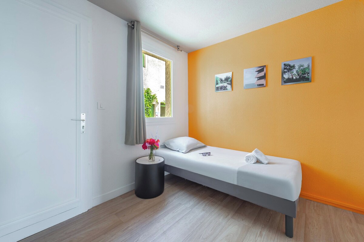 A single-bed room with a window, a bedside table with a vase of flowers, and two wall-mounted pictures.