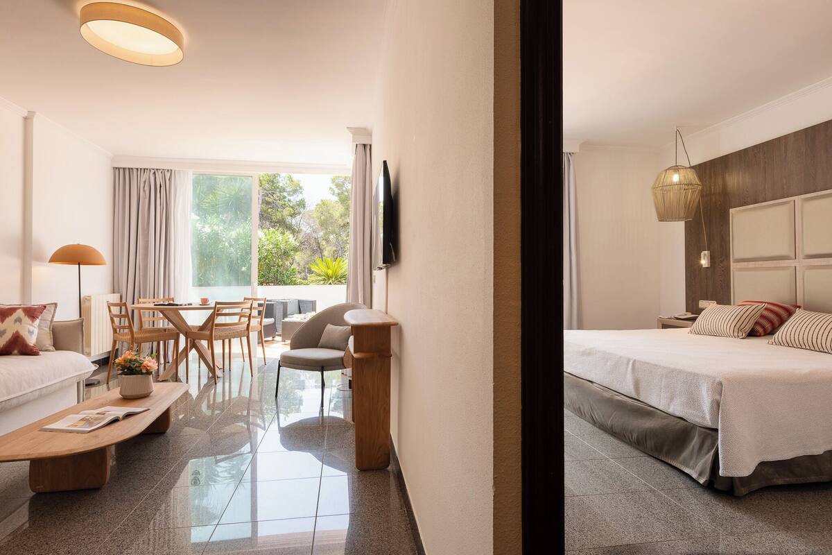 A hotel room with a bed, a sofa, a dining table with chairs, a TV mounted on the wall, and a balcony with curtains.