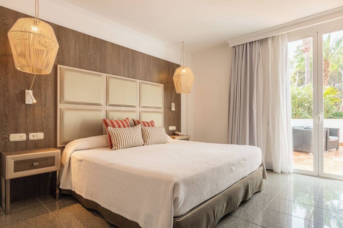 A hotel room with a large bed, two bedside tables, a wall-mounted light fixture, and a view of the outdoors.