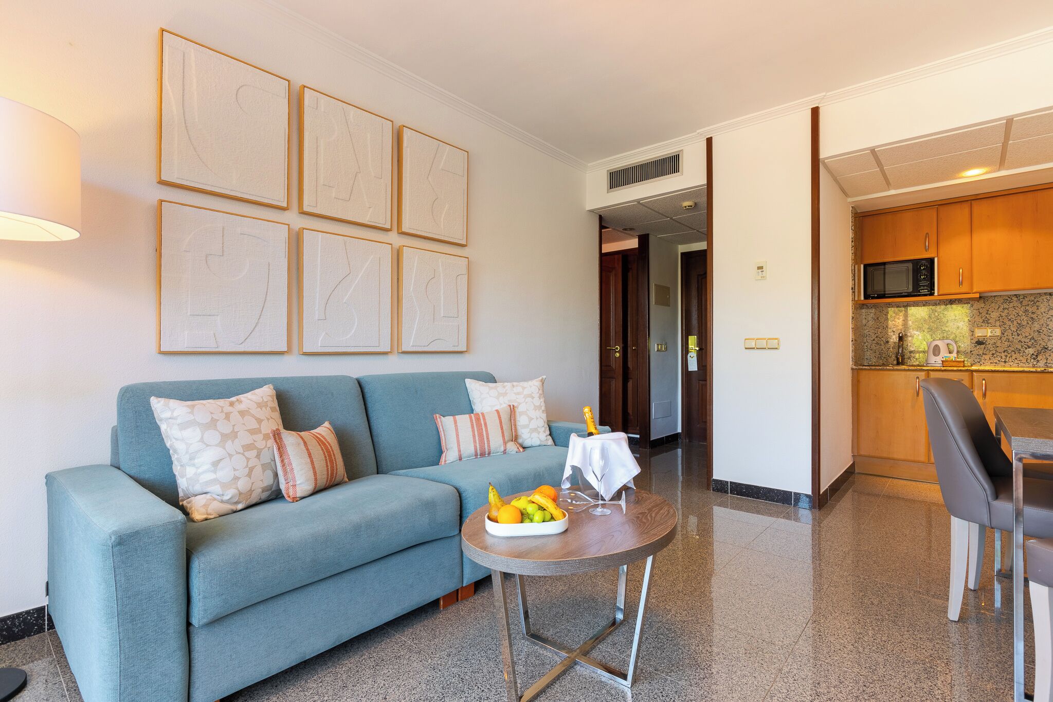 A modern living room with a sofa, a small round table with fruit, and a kitchen in the background.