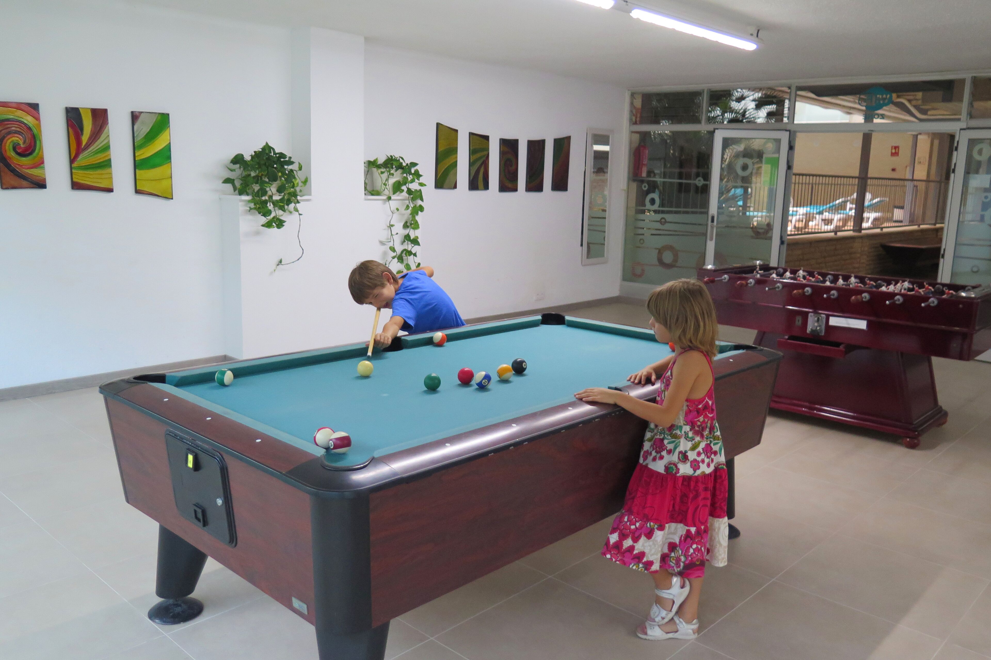 Una mesa de billar con bolas, una mesa de fútbol de mesa y una planta en una habitación con paredes blancas y obras de arte.
