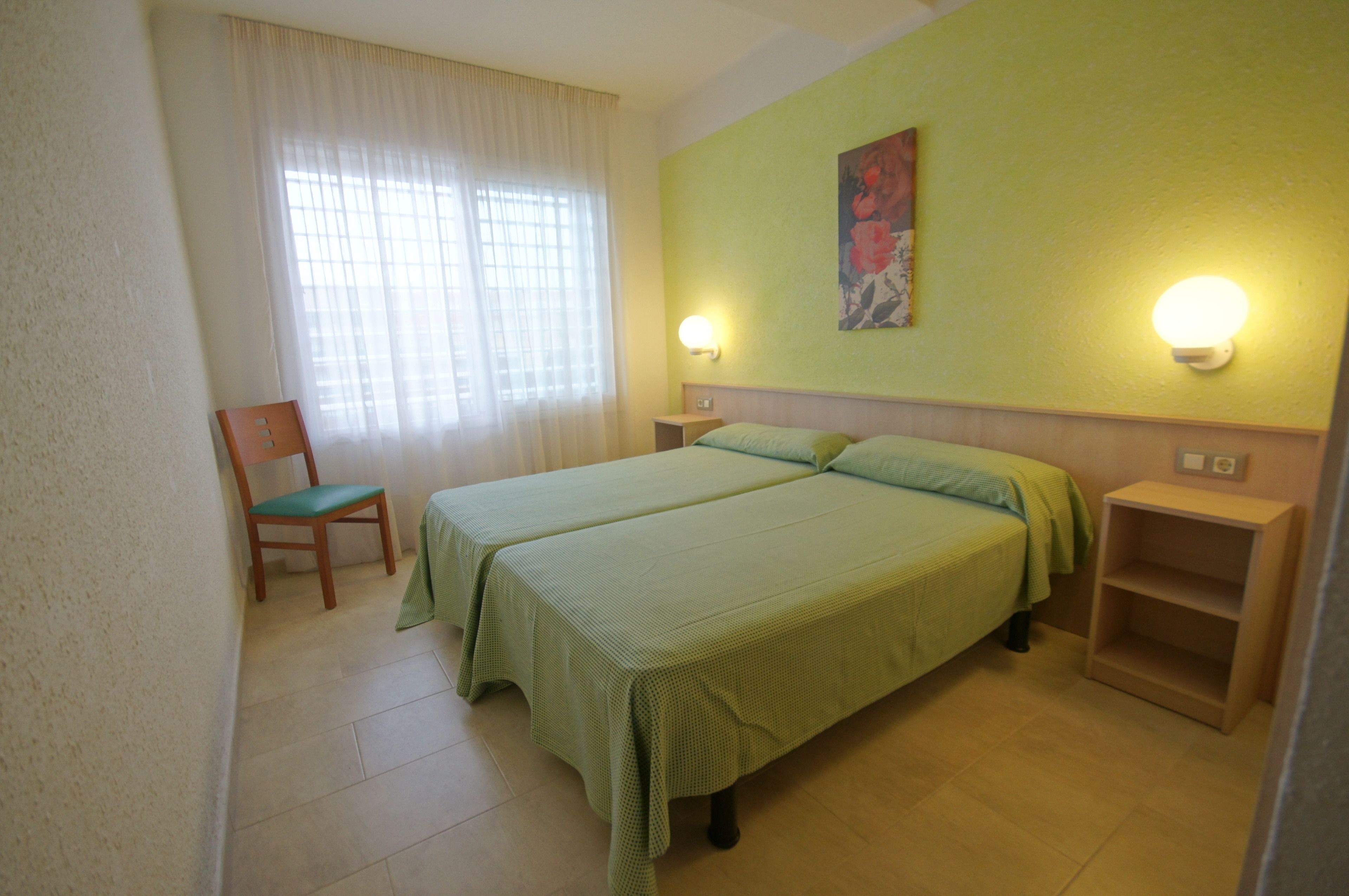 A room with two beds, a chair, a nightstand, and a wall-mounted light fixture.