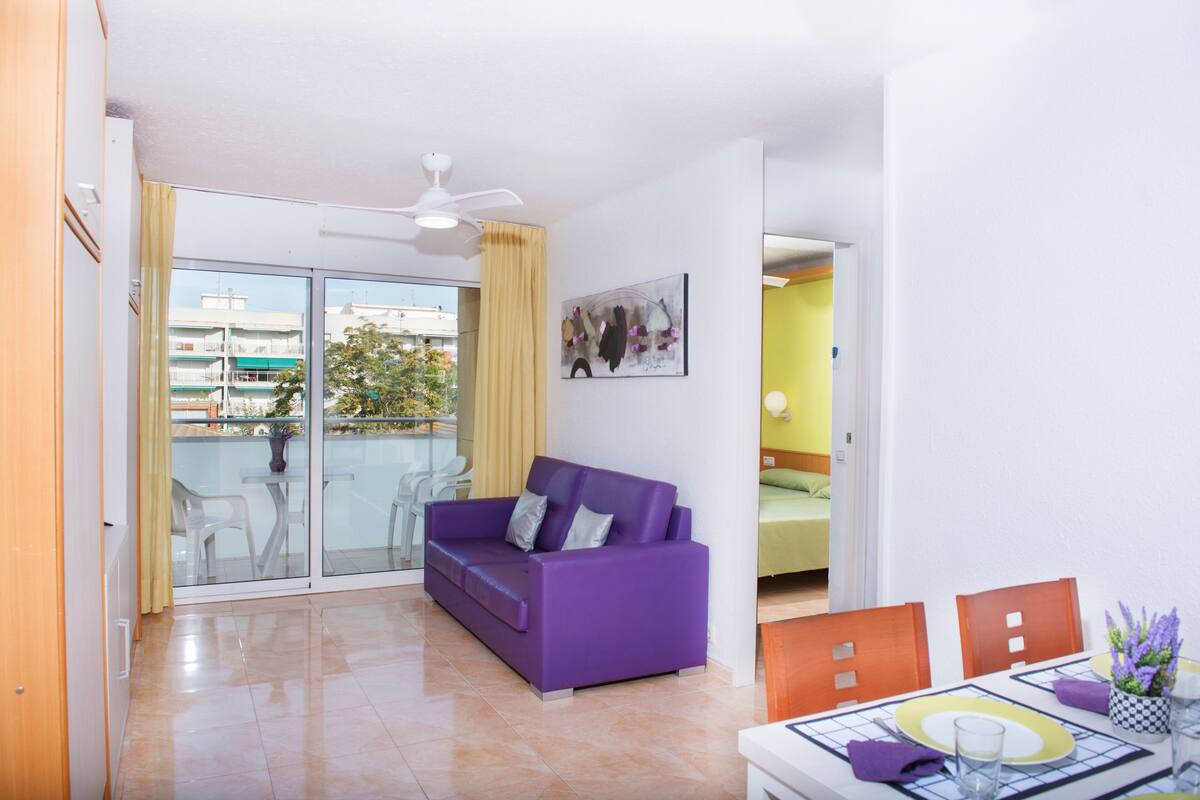 Una sala de estar moderna con un sofá morado, una mesa de comedor con sillas y un balcón con vistas a edificios.