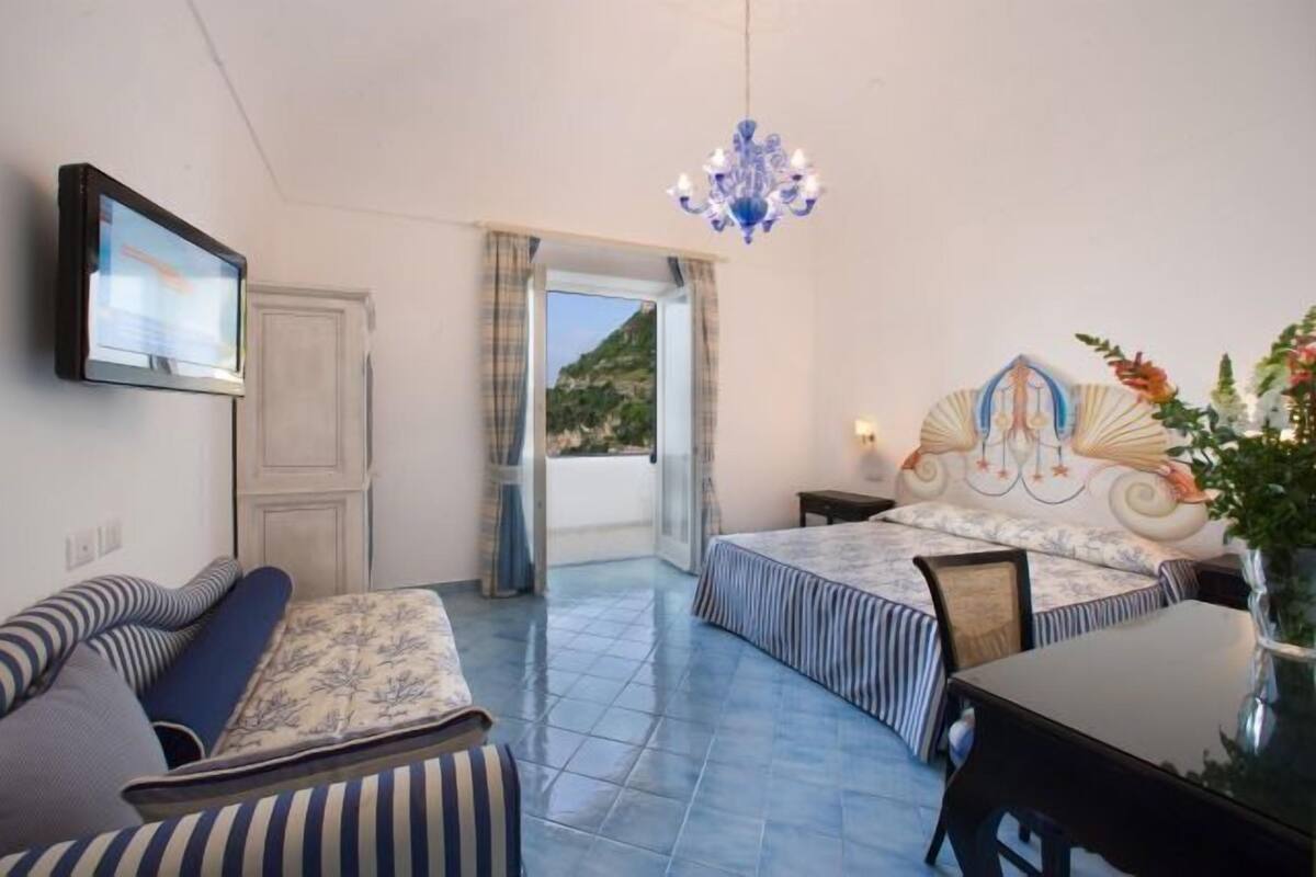 A bedroom with a bed, a sofa, a TV, a chandelier, and a view of the outdoors.