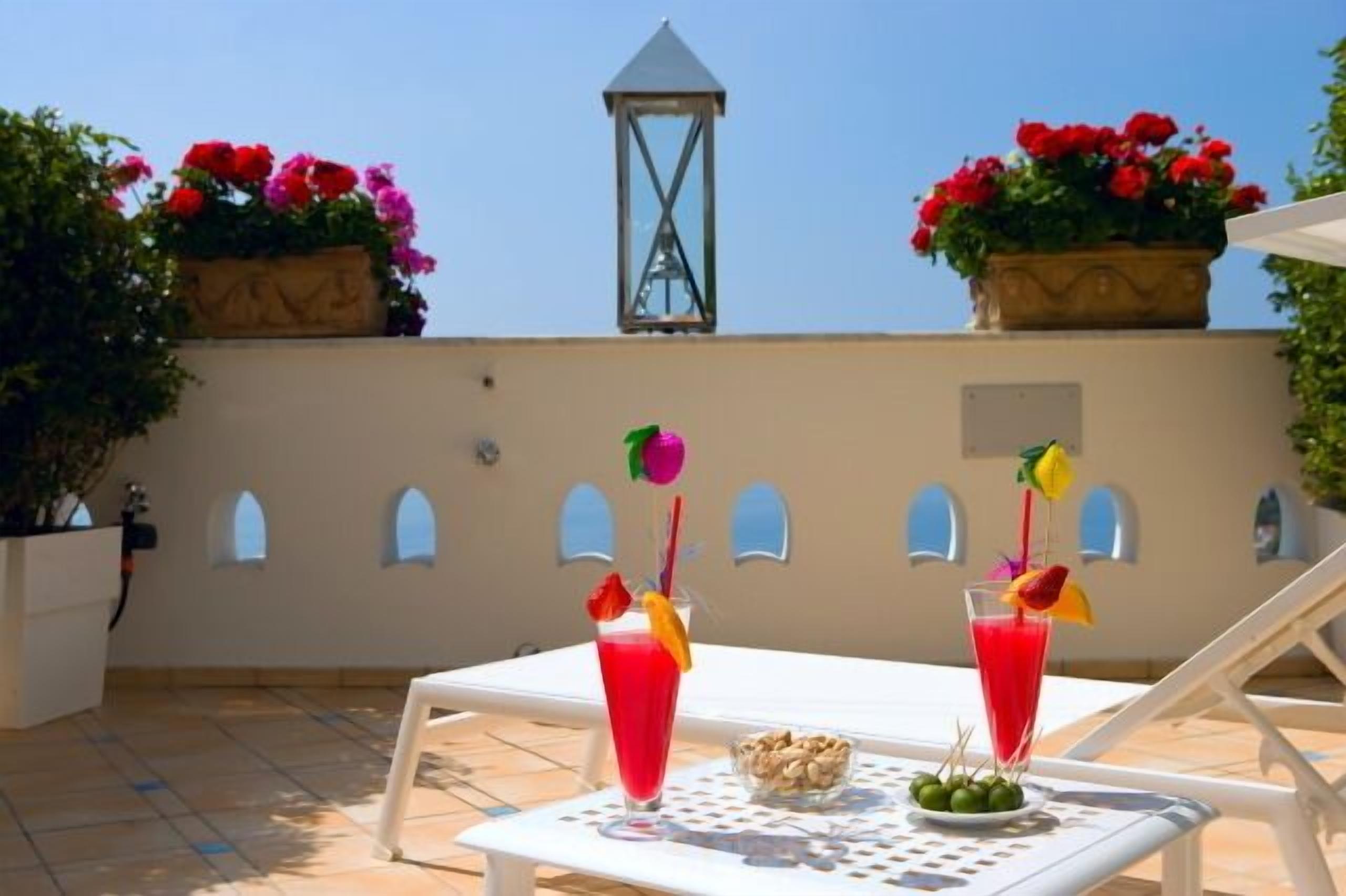 A rooftop terrace with two lounge chairs, a table with a drink and snacks, and flower planters.