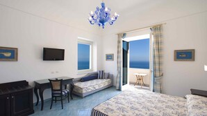 A bedroom with a bed, a desk, a chair, a sofa, a TV, and a view of the sea. - Palazzo Marzoli Resort (Positano)
