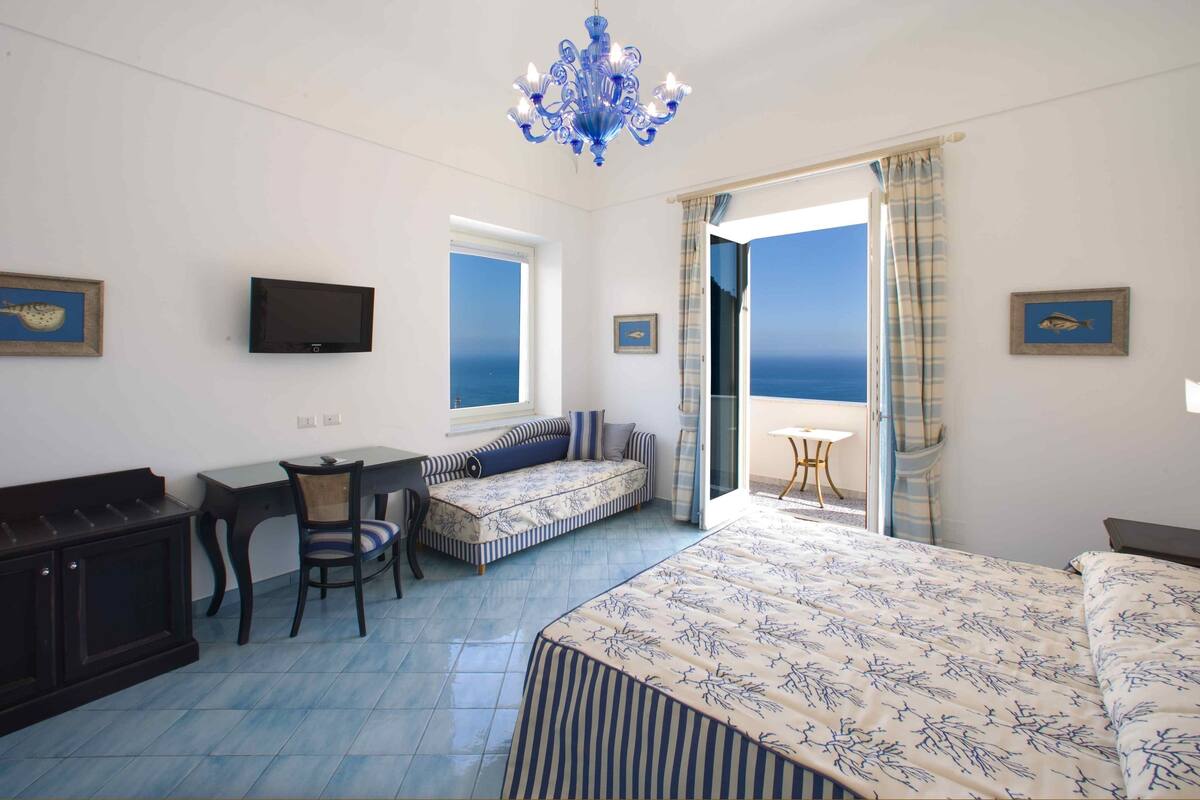 A bedroom with a bed, a desk, a chair, a sofa, a TV, and a view of the sea.