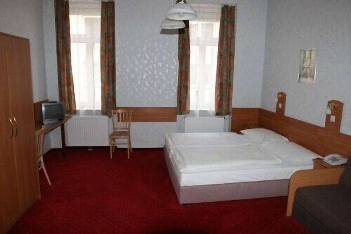 A hotel room with a single bed, a wooden chair, a small desk with a television, and two windows with curtains.