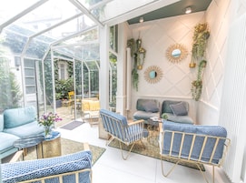 A conservatory with a glass ceiling, furnished with blue and grey sofas, patterned chairs, and a small table.