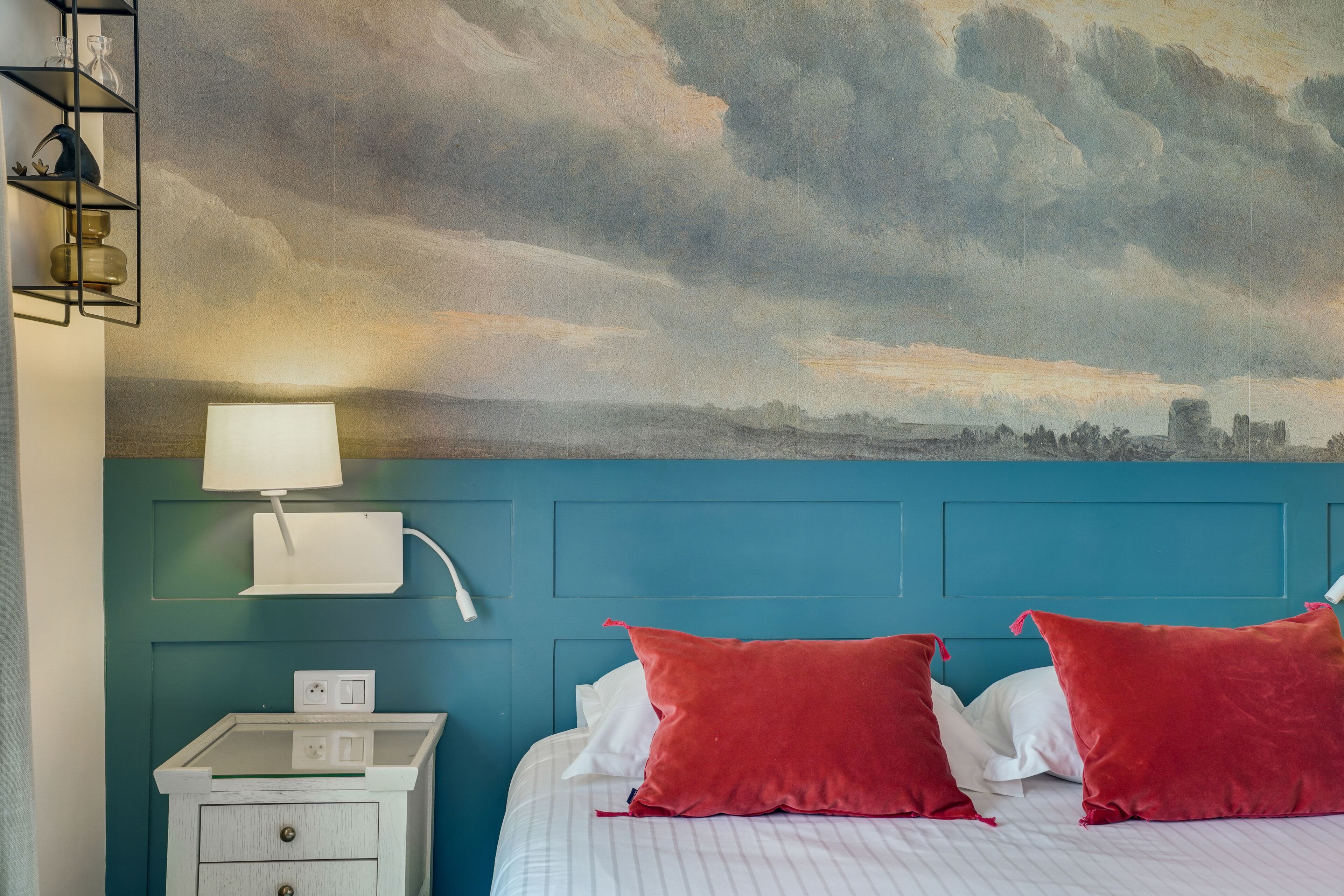 A hotel room with a bed, a nightstand, a lamp, and a wall mural of a cloudy sky.