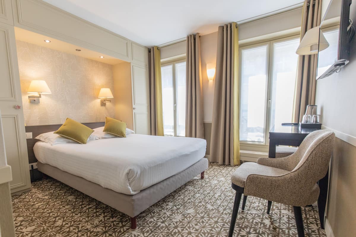 A hotel room with a large bed, two bedside lamps, a chair, a desk with a lamp, and patterned carpet.