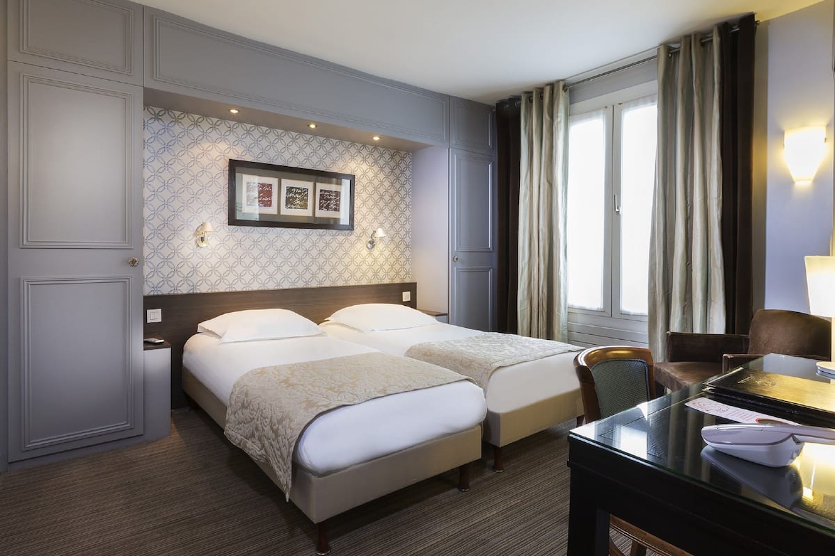 A hotel room with two beds, a desk with a lamp, a chair, and a window with curtains.