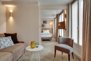Habitación de hotel con sofá, sillón y una mesita con un jarrón y vaso. Se ve una cama con cabecera y una ventana con cortinas.