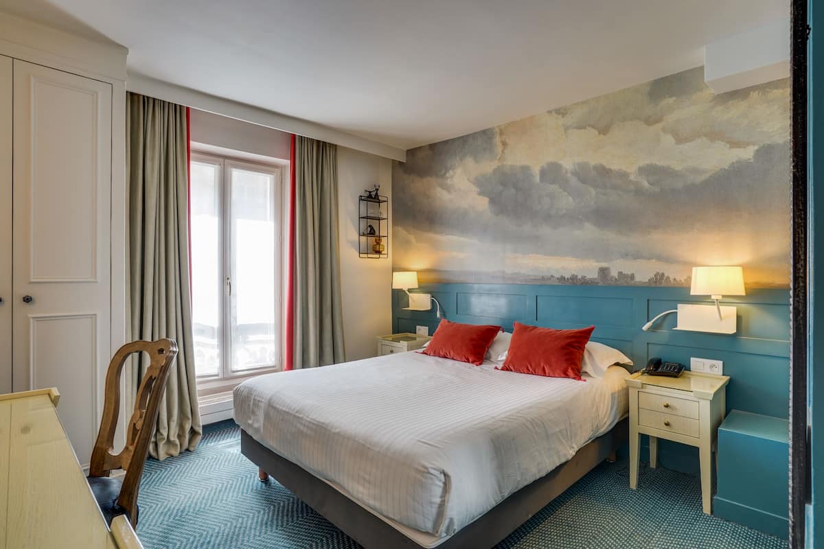 A hotel room with a bed, two bedside tables, a chair, a view of the city, and a wall mural.
