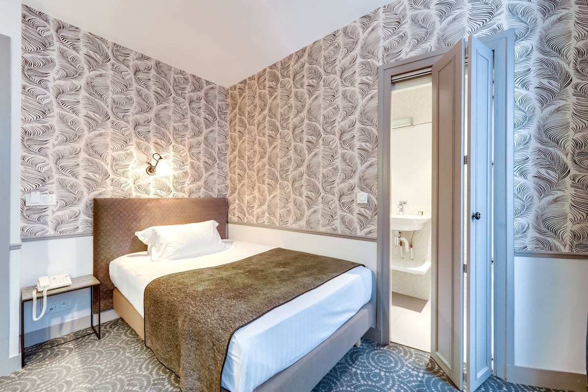 A hotel room with a patterned wallpaper, a bed with a brown blanket, a bedside table with a phone, and a bathroom visible through an open door.