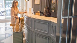 A reception desk with a grey panel and a framed menu. A woman is standing at the counter, and a suitcase is visible in the foreground. - Hotel Passy Eiffel (Paris)
