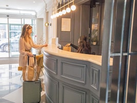 A reception desk with a grey panel and a framed menu. A woman is standing at the counter, and a suitcase is visible in the foreground.