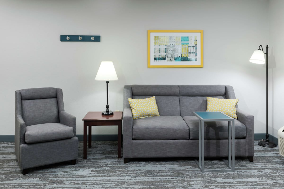 A room with a grey sofa, a grey armchair, a small table, a floor lamp, and a framed abstract painting on the wall.