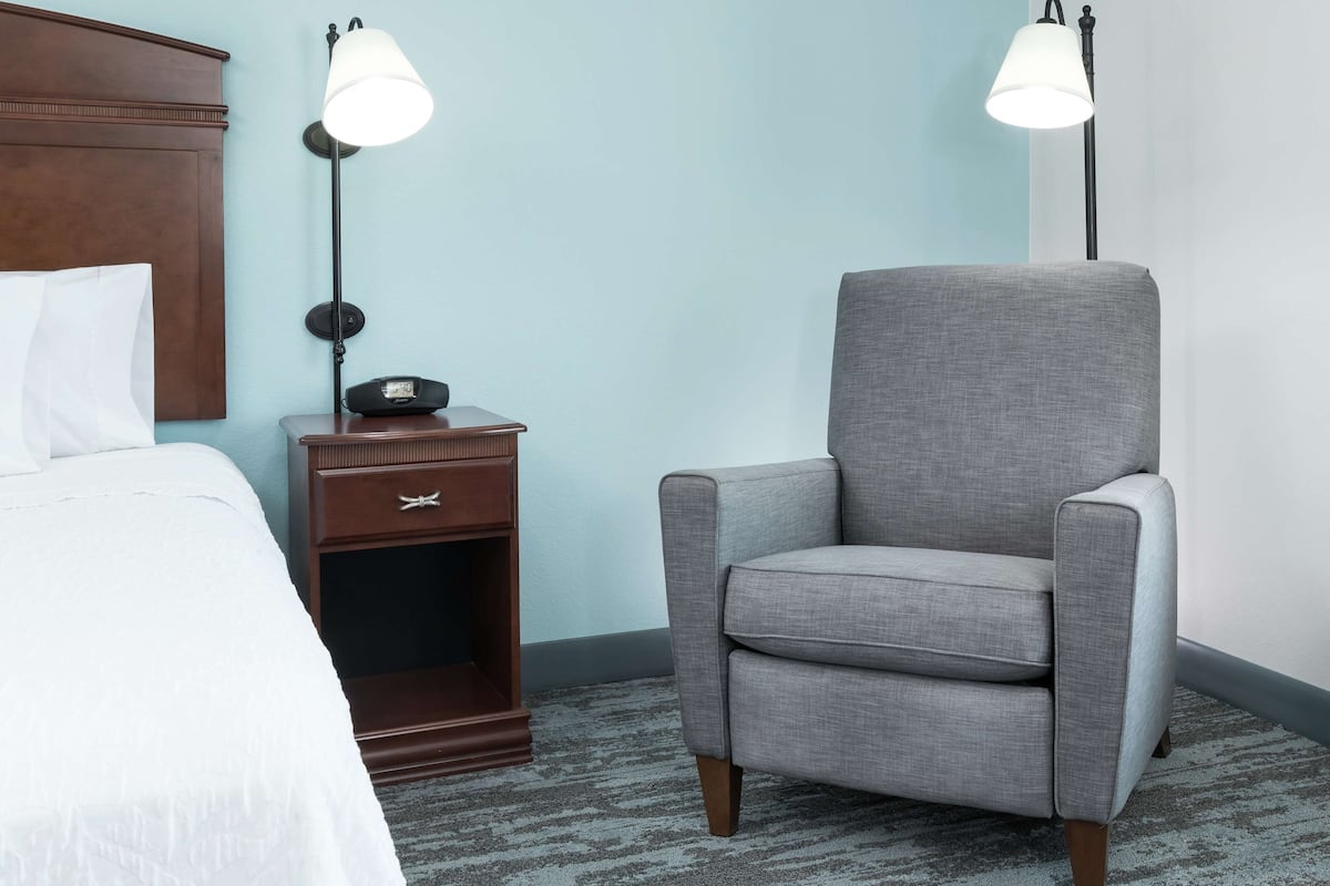 A hotel room with a bed, a nightstand with a lamp, a chair, and a clock.