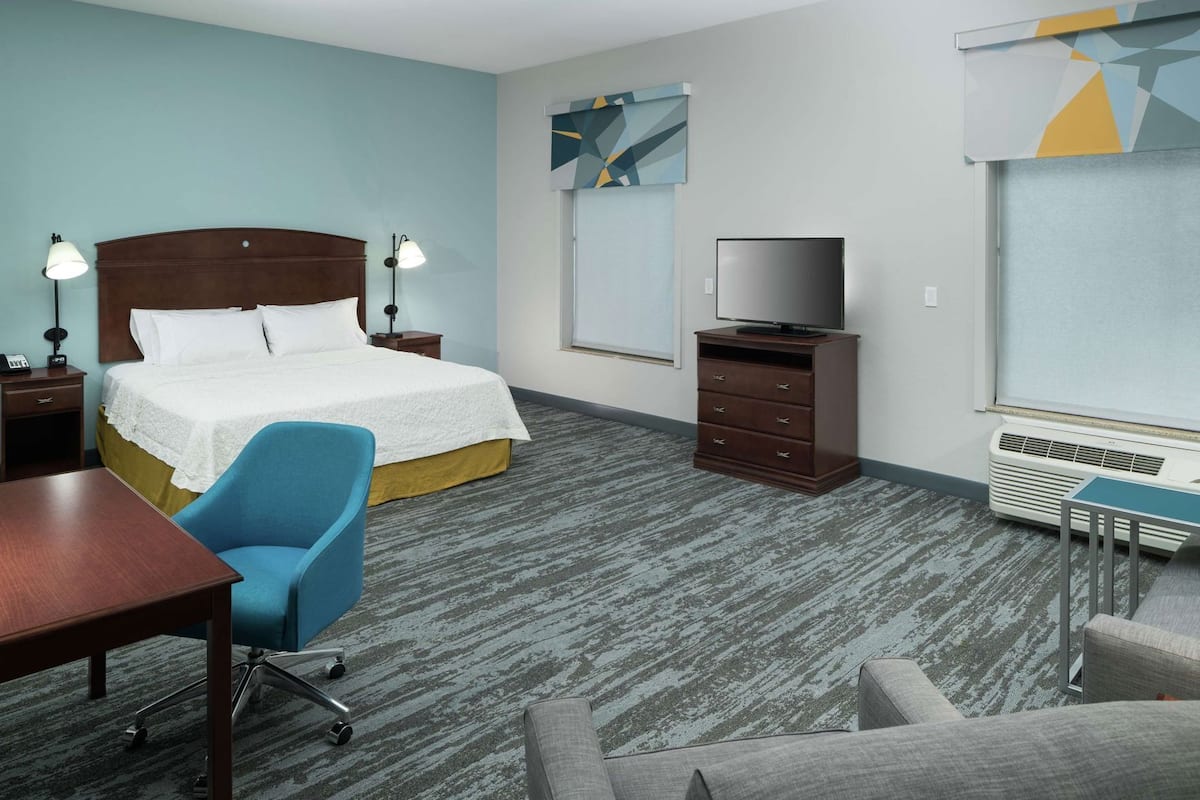A hotel room with a bed, a desk, a chair, a TV, and a heater.