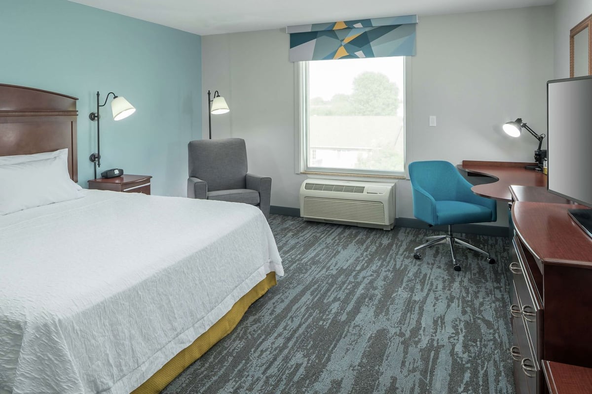 A hotel room with a bed, a desk, a chair, a desk lamp, a standing lamp, and a window with a view.