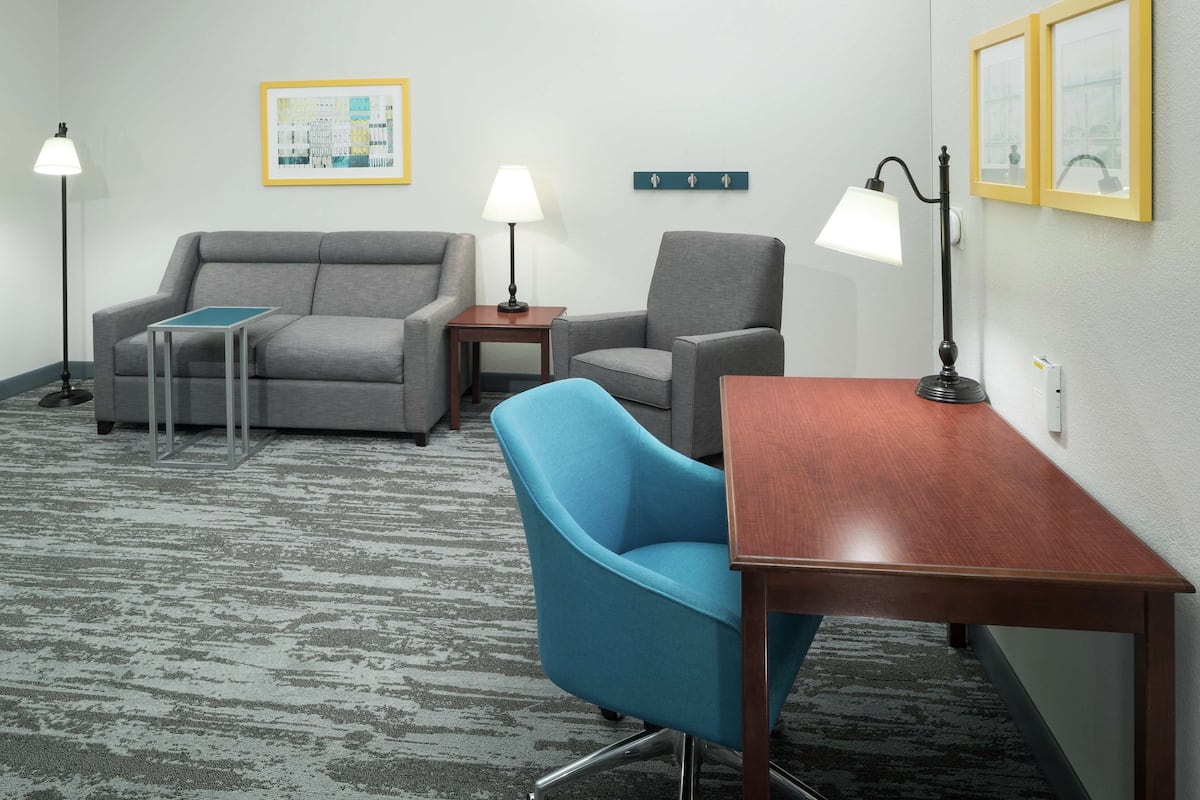 A hotel room with a gray sofa, a blue armchair, a wooden table, two floor lamps, a framed picture on the wall, and a carpeted floor.