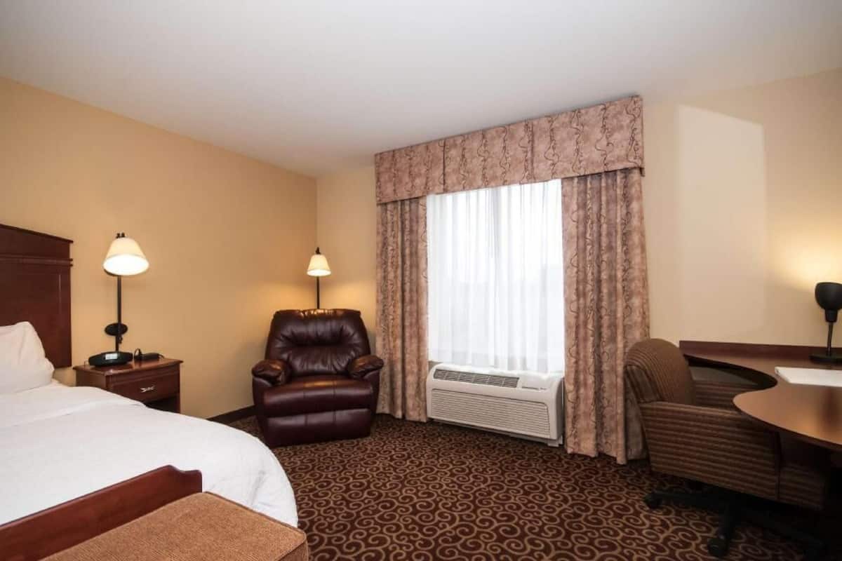A hotel room with a bed, two chairs, a desk, a lamp, a window with curtains, and a patterned carpet.