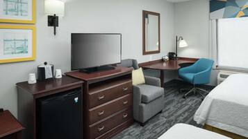 A hotel room with a flat-screen TV, a desk with a chair, a small refrigerator, a bedside table with a lamp, and a bed.