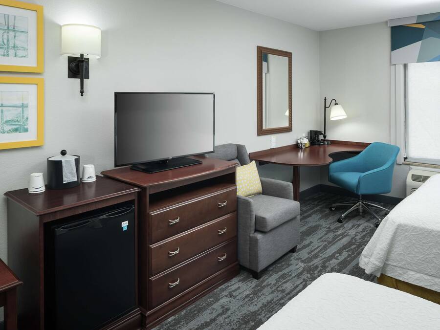 A hotel room with a flat-screen TV, a desk with a chair, a small refrigerator, a bedside table with a lamp, and a bed.