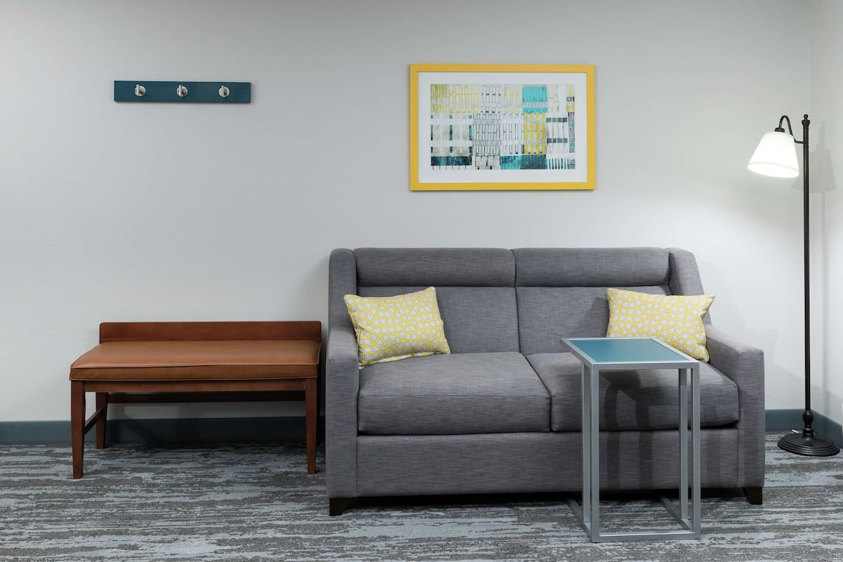 A grey sofa with two pillows, a small table, a bench, a framed picture, and a floor lamp.