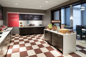A modern kitchen with checkered flooring, a central island with food items, and a sign indicating breakfast starts here.