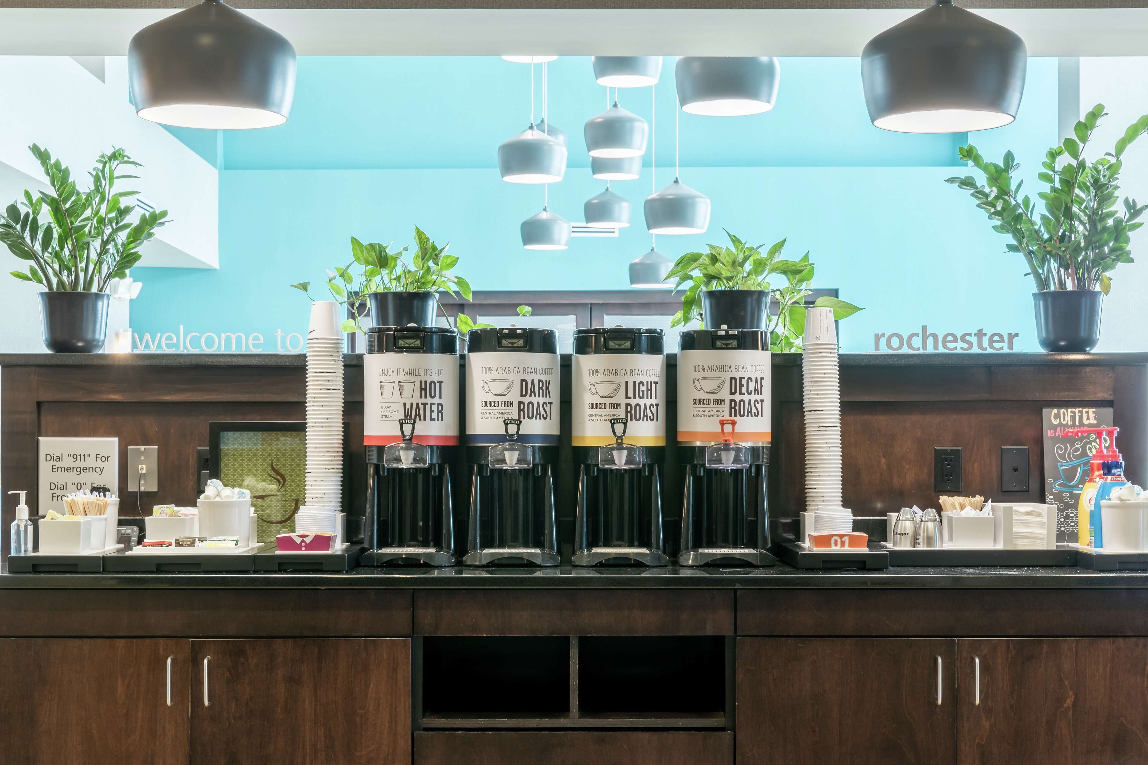 A coffee station with four dispensers labeled "HOT WATER," "DARK ROAST," "LIGHT ROAST," and "DECAF ROAST," each with a coffee cup and spoon.