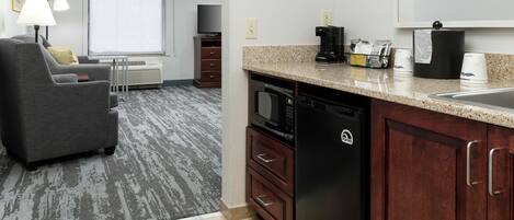 A hotel room with a gray sofa, a small dining table, a microwave, a dishwasher, a coffee maker, and a large window with blinds.