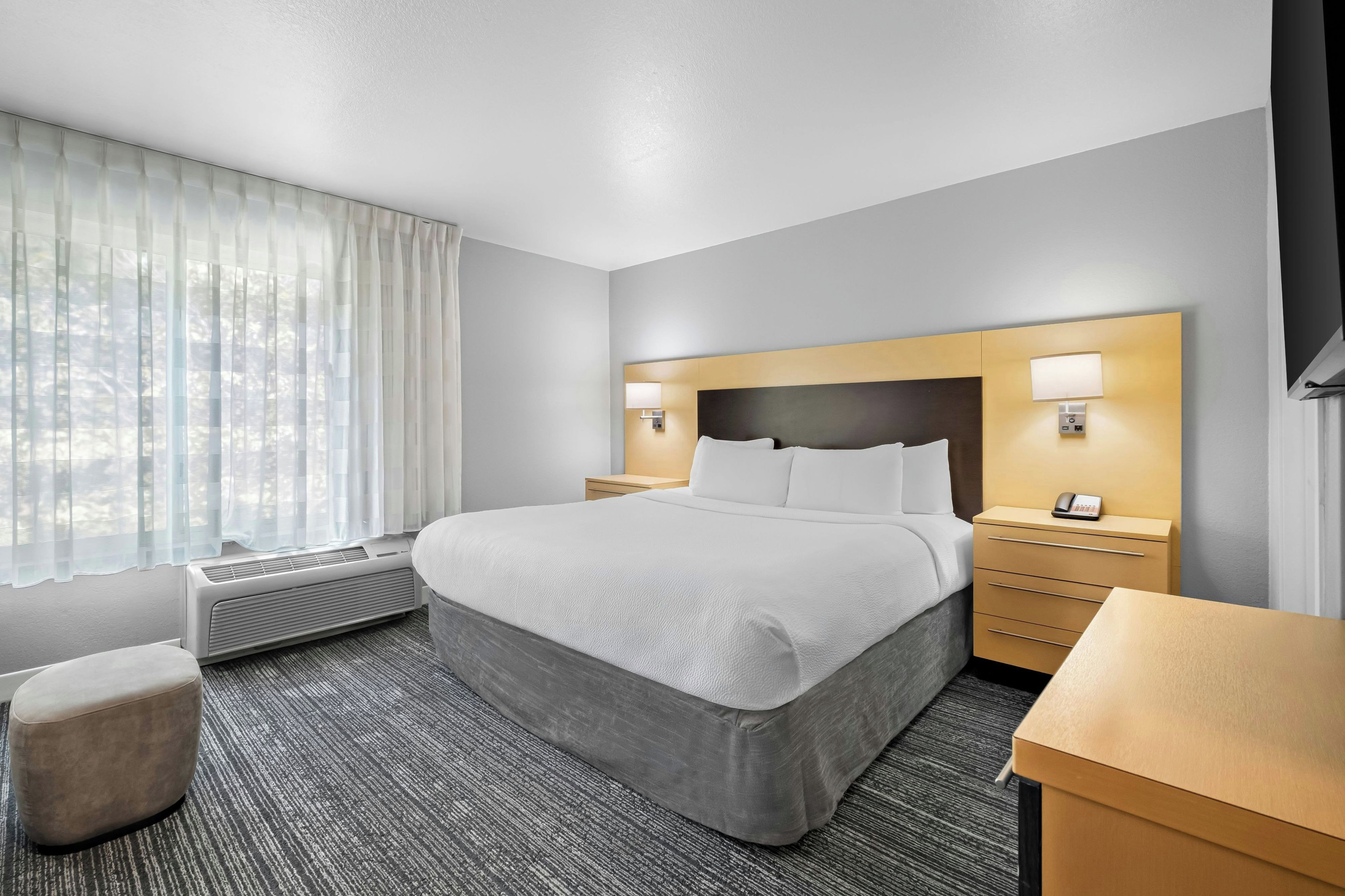 Quarto de hotel com cama, mesas de cabeceira, escrivaninha, cadeira, janela com cortinas e unidade de ar condicionado.