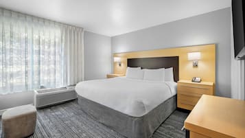 Quarto de hotel com cama, mesas de cabeceira, escrivaninha, cadeira, janela com cortinas e unidade de ar condicionado.