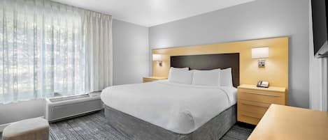 A hotel room with a bed, bedside tables, a desk, a chair, a window with curtains, and an air conditioning unit.