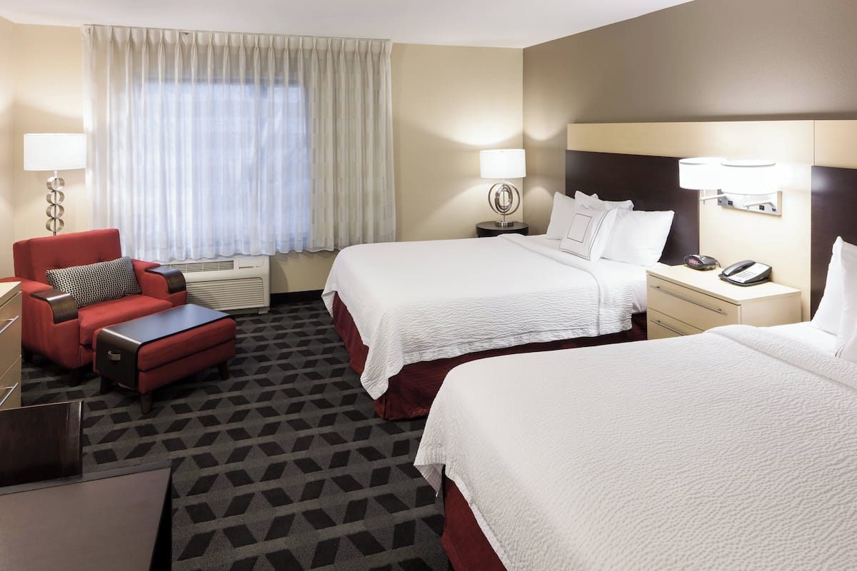 Une chambre d’hôtel avec deux lits, un fauteuil rouge, une petite table et une table de chevet avec un téléphone et une lampe.