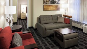 A hotel room with a grey sofa, red armchair, and a small table. There is a painting on the wall and a lamp on a side table. - TownePlace Suites by Marriott Little Rock West (Little Rock)
