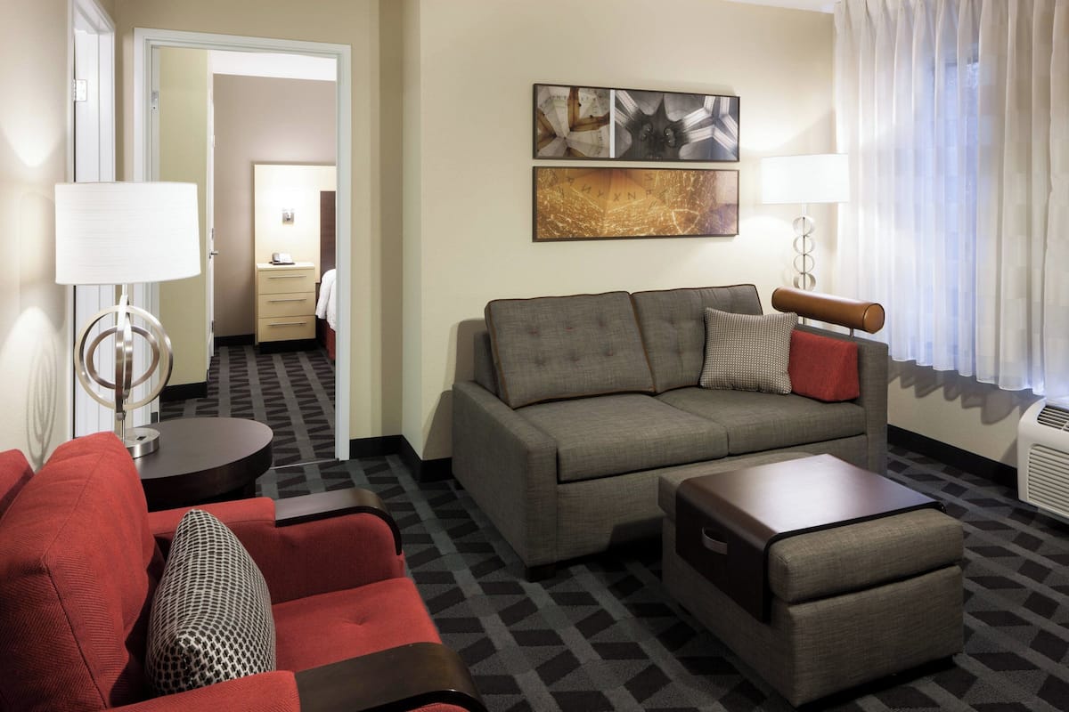 Une chambre d’hôtel avec un canapé gris, un fauteuil rouge et une petite table. Il y a un tableau au mur et une lampe sur une table d’appoint.