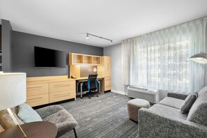 A modern hotel room with a flat-screen TV, a desk with a chair, a sofa, and a large window with sheer curtains.